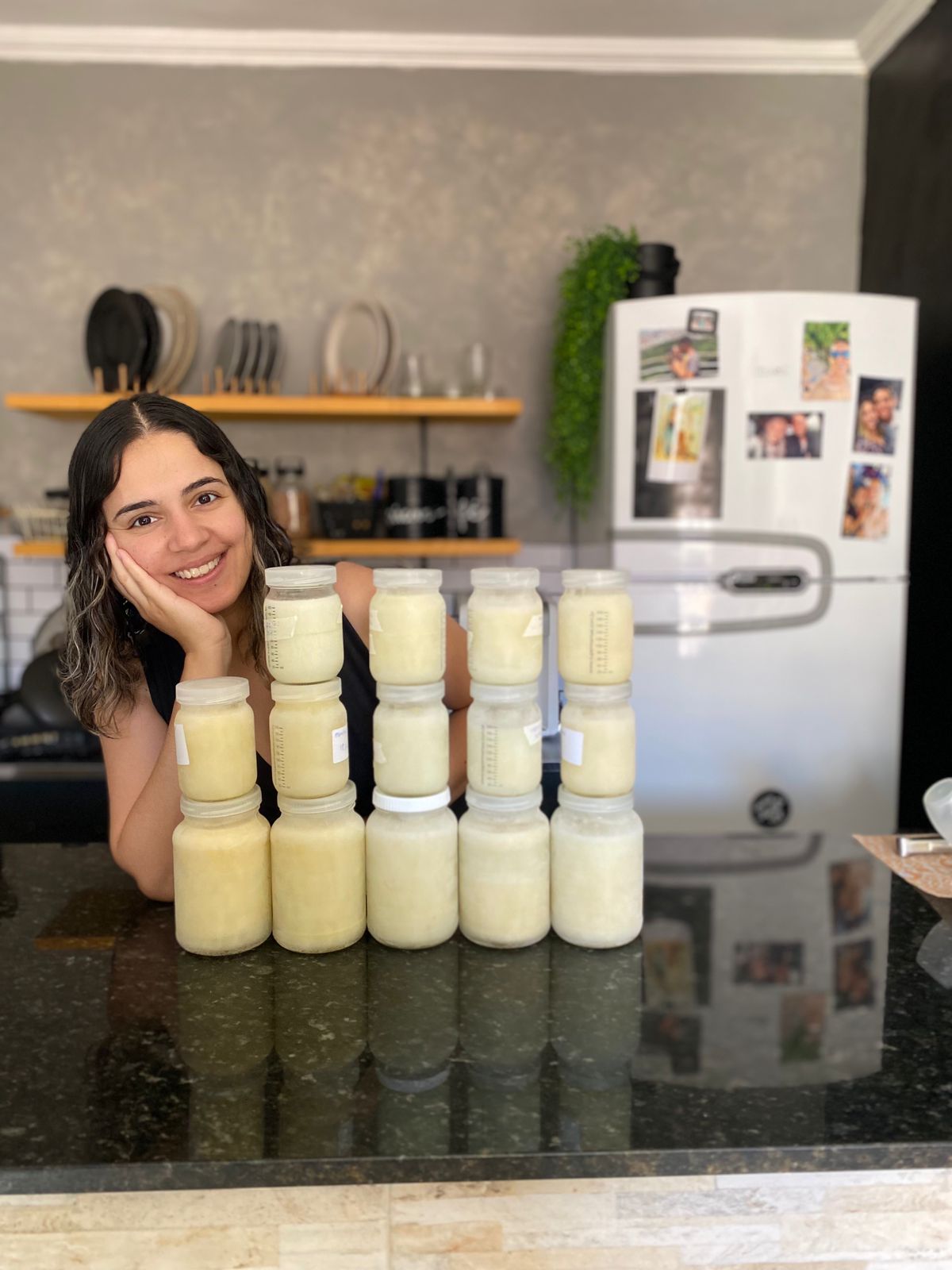 Na imagem, Maniely, uma mulher branca e de cabelos castanhos, está em uma cozinha. Posicionado ao lado esquerdo da imagem, está apoiada sobre um balcão com a mão no queixo, e ao seu lado há  14 potes com leite humano empilhados em três fileiras. Ao fundo, há uma parede cinza, alguns pratos, copos e uma geladeira, com um pequeno vaso sobre ela.